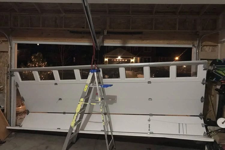 Sectional garage door pulled off its tracks and leaning inward, propped on a stepladder during a night-time emergency call, with the street lit beyond the open doorway