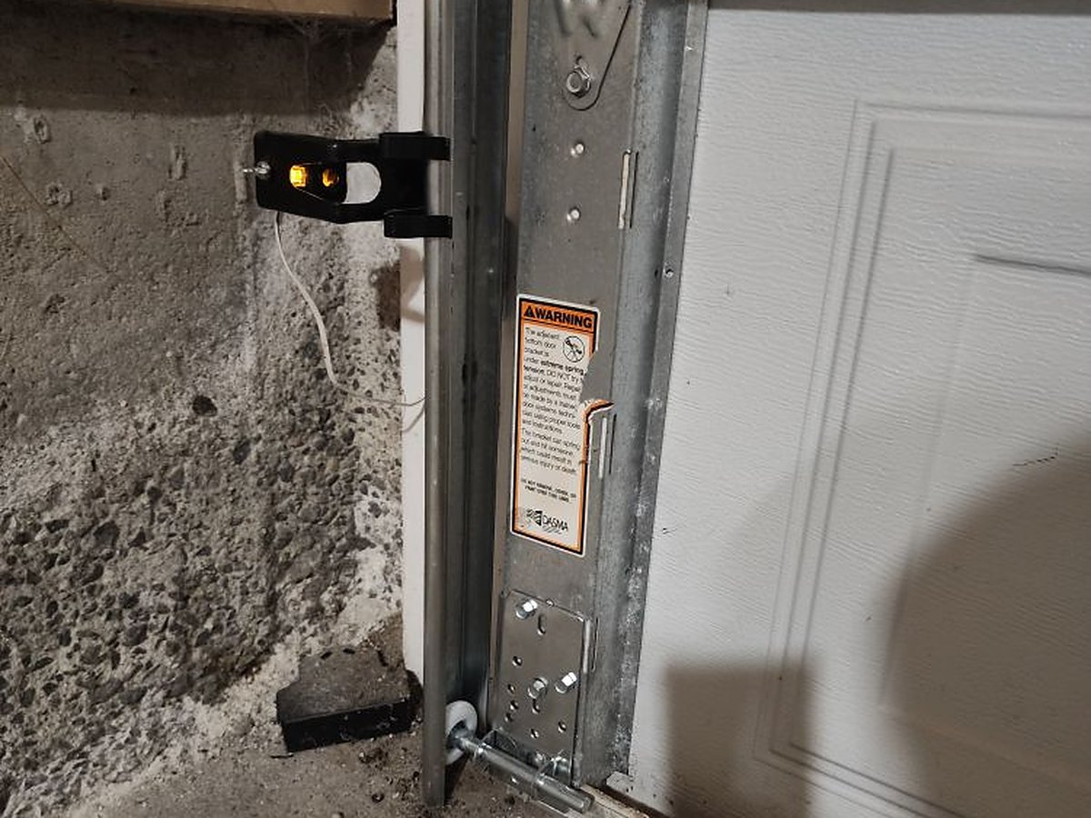 Vertical track, bottom roller bracket and lift cable at the base of a garage door, with the wall-mounted safety sensor beside the opening